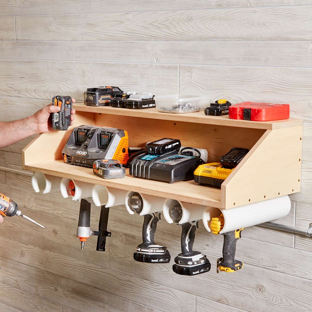 A hand organizes various power tools and chargers on a wooden wall-mounted shelf in a well-lit workshop with a textured wooden backdrop.