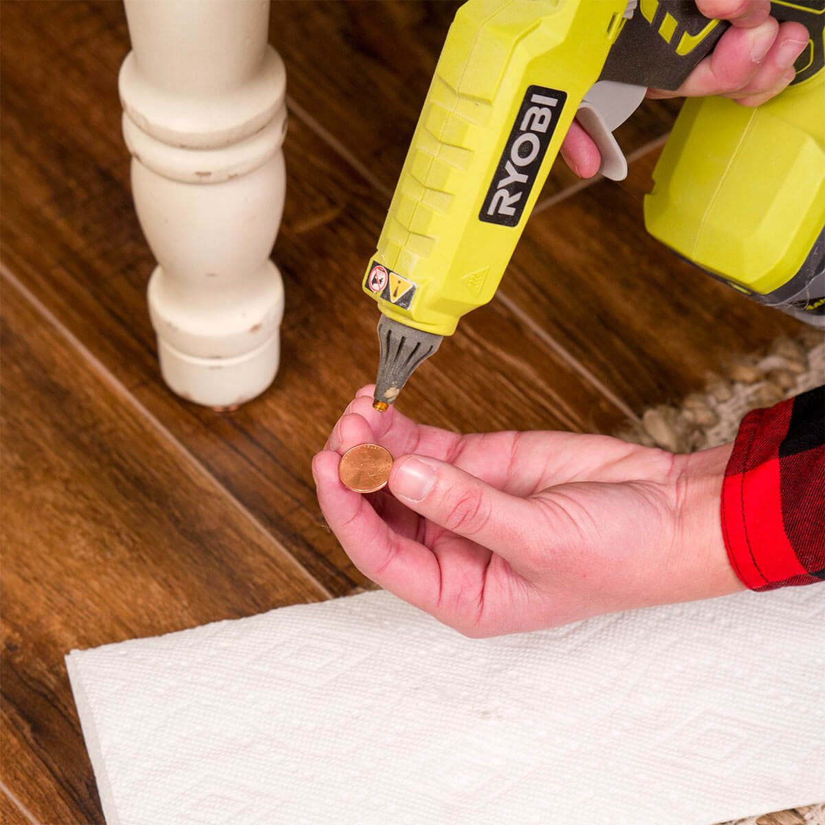 A person holds a penny while using a hot glue gun above it. The setting features wooden flooring and a nearby paper towel.