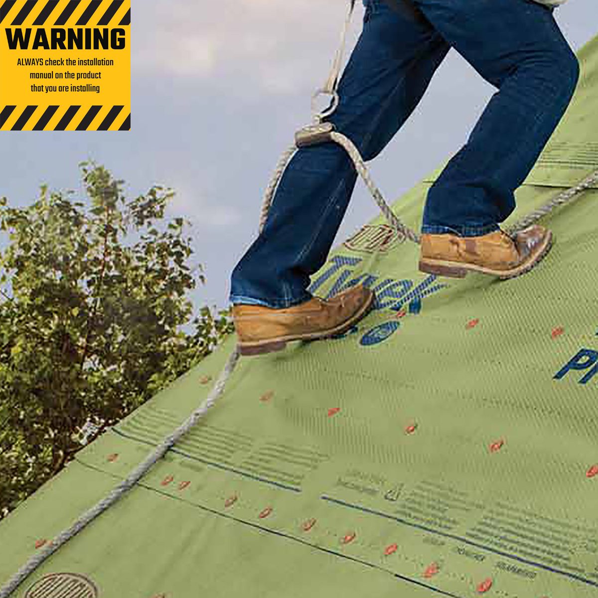 A person is standing on a sloped green roof surface, secured with a harness and rope, while trees and a cloudy sky are in the background.