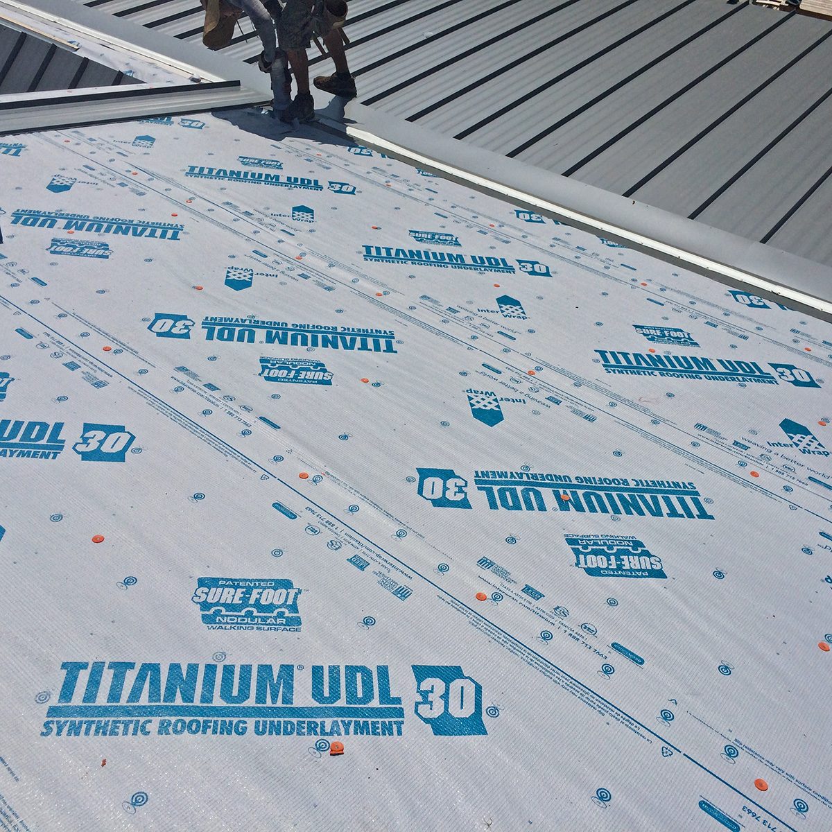 Synthetic roofing underlayment is being installed on a sloped roof, with workers positioned above, surrounded by metal roofing sheets in a bright outdoor setting.