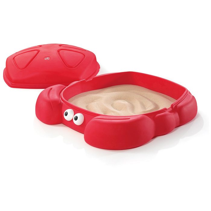 A red crab-shaped sandpit sits open, revealing light-colored sand inside, accompanied by a matching lid nearby, creating a playful outdoor activity space.