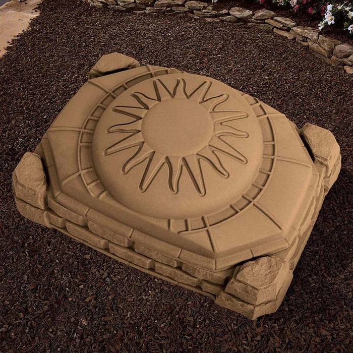A large, sandy-colored, sun-patterned stone sits atop dark mulch, surrounded by a rocky border and flowering plants in a garden setting.