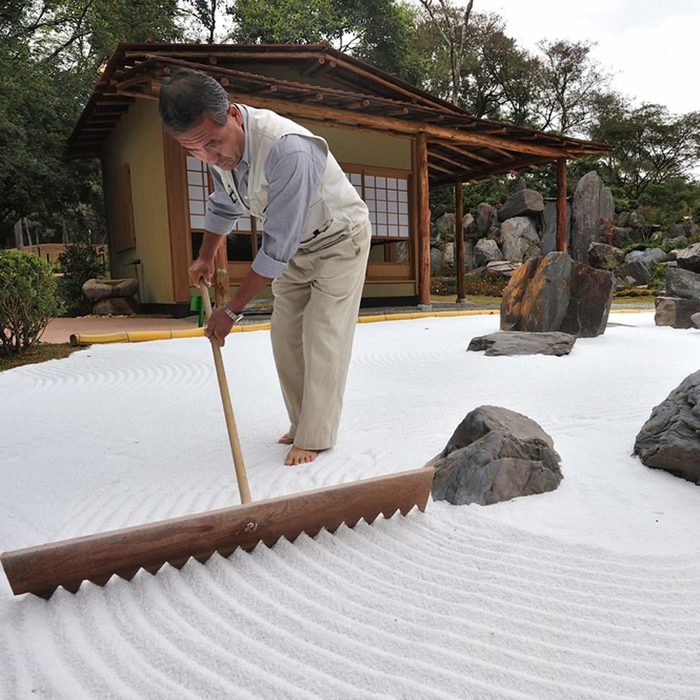 A man uses a rake to create patterns in white sand, surrounded by stones and a traditional wooden building in a garden setting.