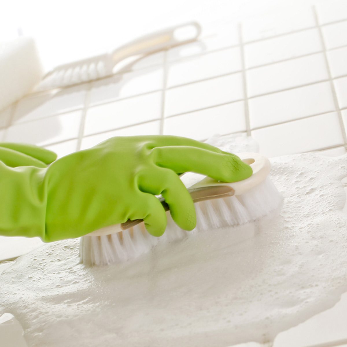 A hand in a green glove scrubs a soapy surface with a brush, surrounded by a clean, tiled environment.