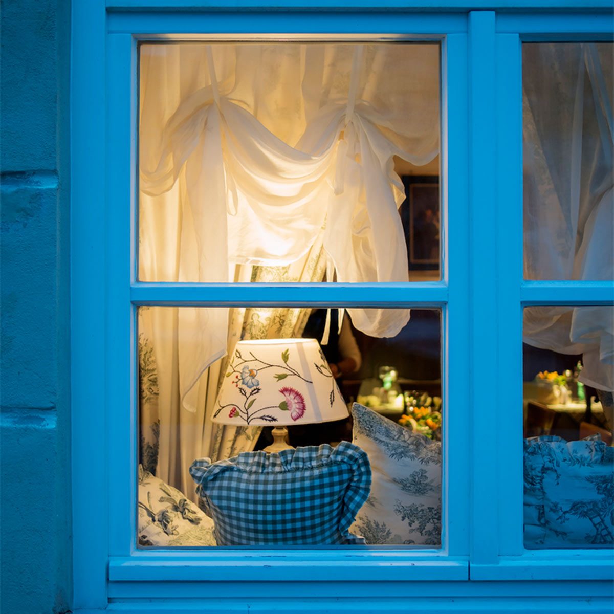 A lamp illuminates a cozy interior, showcasing cushions and draped curtains, viewed through a blue-framed window in a warmly lit setting.