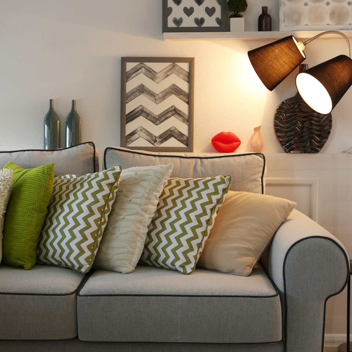 A beige couch adorned with patterned pillows sits under a wall lamp, alongside decorative objects and framed art, creating a cozy living space.