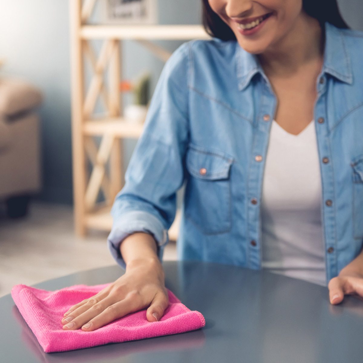 A woman wipes a table with a pink cloth, smiling, in a cozy room with wooden shelves and soft furnishings in the background.