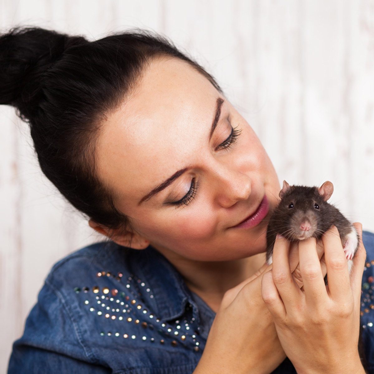 A woman gently holds a rat close to her face, smiling affectionately. The background features soft, light-colored wood, suggesting a cozy indoor setting.