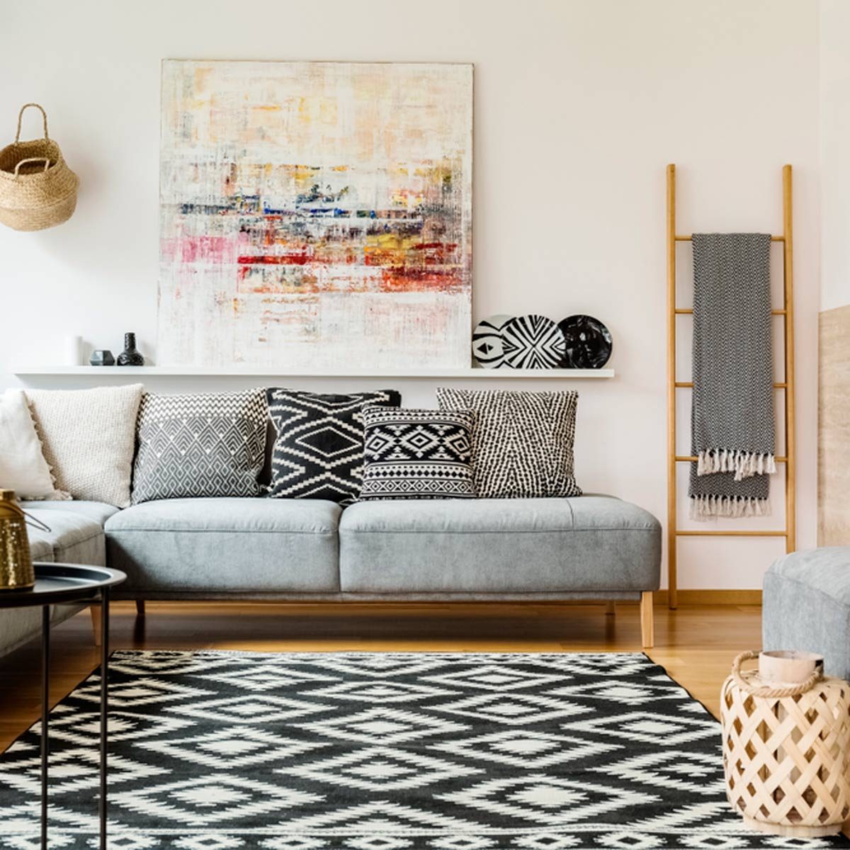 A gray sofa with patterned pillows rests on a black and white rug. Colorful art hangs above, surrounded by decorative items and a woven storage basket.