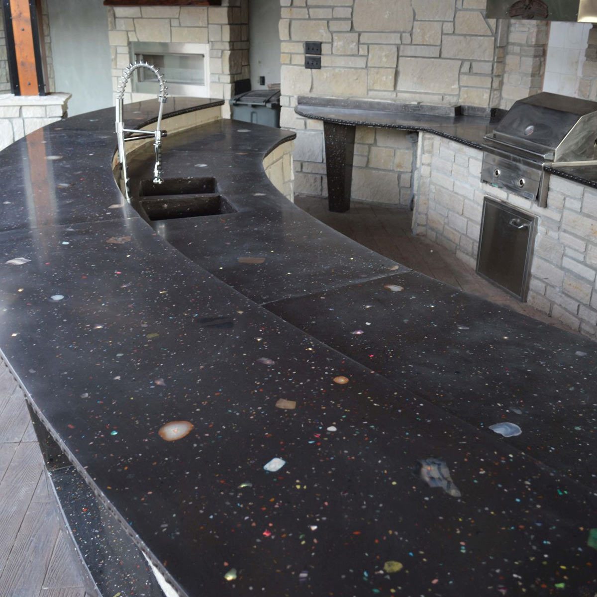 A curved black countertop with multicolored speckles sits in a stone-walled outdoor kitchen, featuring a faucet and appliances in the background.