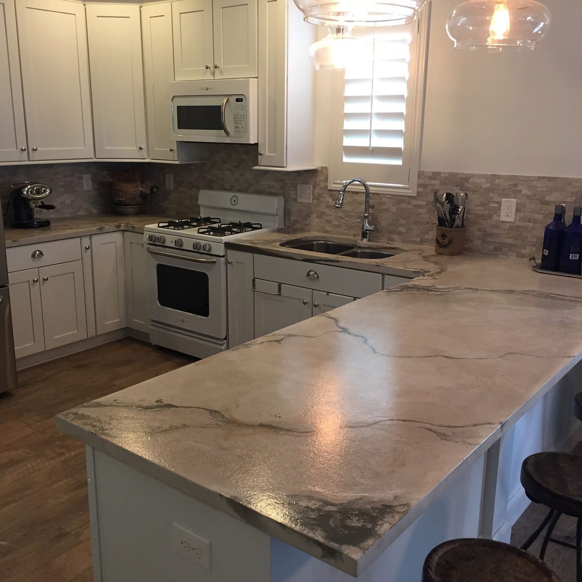 A kitchen features white cabinets and modern appliances. A countertop with a marble-like texture extends into a seating area, illuminated by hanging lights.