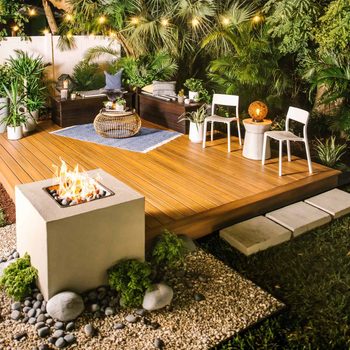 A fire pit burns brightly on a wooden deck, surrounded by greenery and decorative furniture, creating a cozy outdoor gathering space at night.
