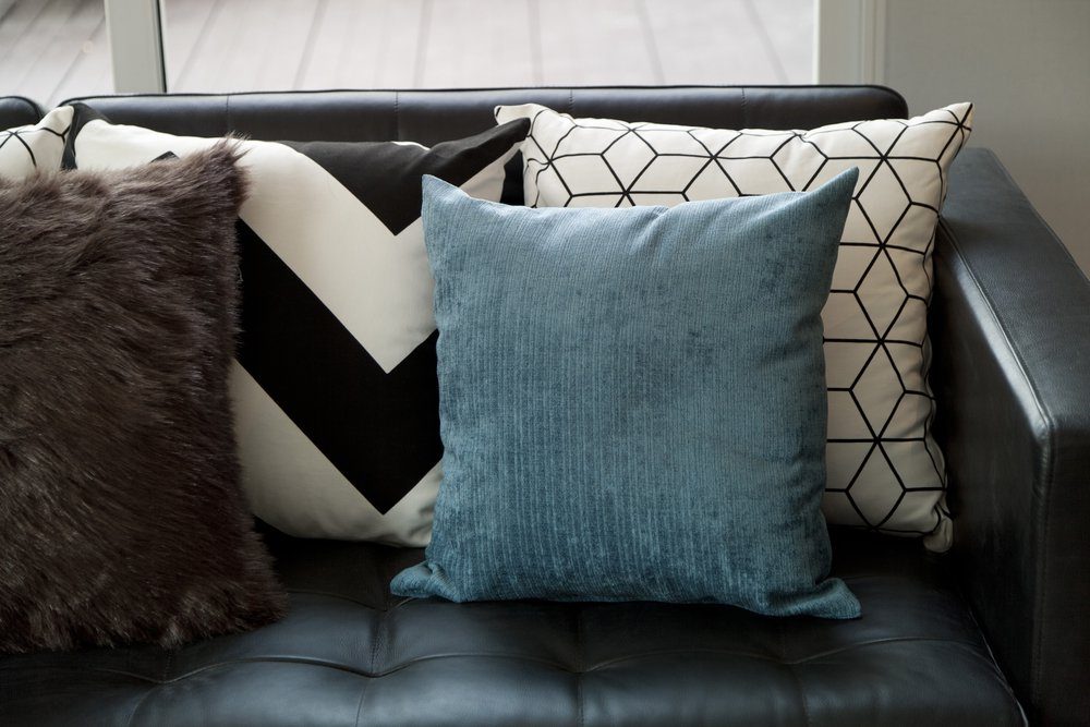 A blue textured pillow rests among various patterned cushions on a black leather sofa, with natural light illuminating the setting.