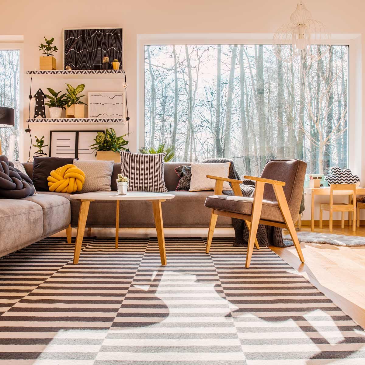 A cozy living room features a gray sofa with various cushions, a round coffee table, and a wooden armchair, illuminated by natural light through large windows.