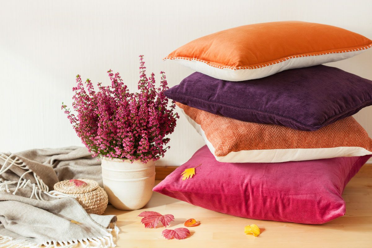 A stack of colorful cushions rests beside a pot of pink flowers, surrounded by a soft blanket and scattered autumn leaves on a wooden surface.