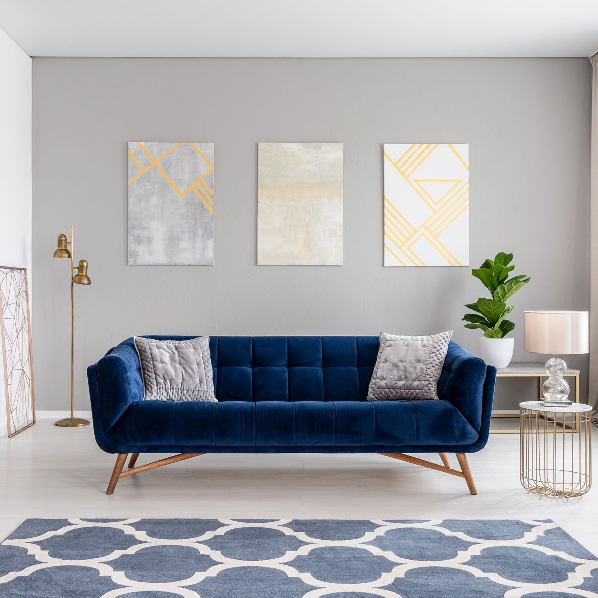 A blue velvet sofa with cushions sits on a patterned rug, beside a plant and decorative lamp, against a gray wall with abstract art.