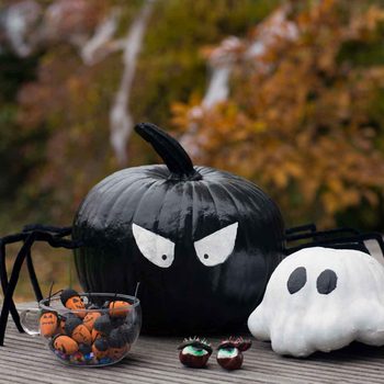 A black pumpkin with spider legs and menacing eyes sits beside a white ghost figure, while a bowl of decorated candy is in front, set outdoors.