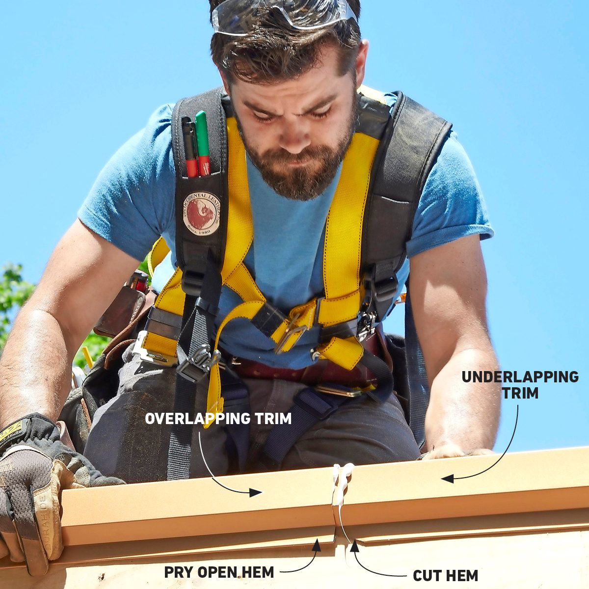 A man in safety gear is working on a roof, focusing on trim details, while surrounded by a clear blue sky and greenery in the background.
