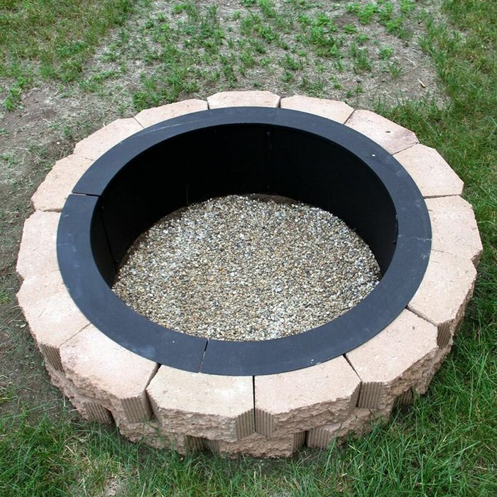 A circular fire pit made of stacked stones holds gravel inside, surrounded by grass in an outdoor setting.