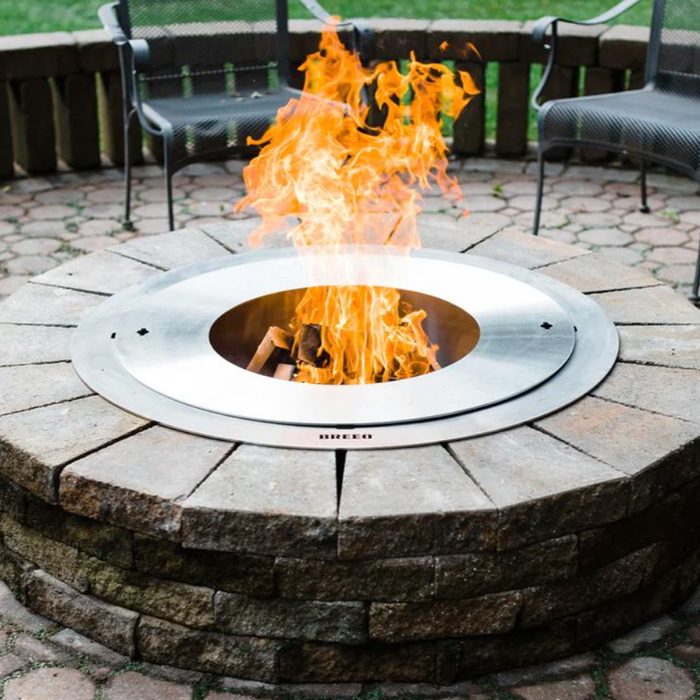 A circular fire pit emits bright flames, surrounded by stone blocks, with two metal chairs positioned nearby on a paved surface in a green outdoor setting.