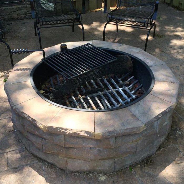 A circular stone fire pit with a metal grate above burning logs rests on a stone patio, surrounded by chairs in a shaded outdoor area.
