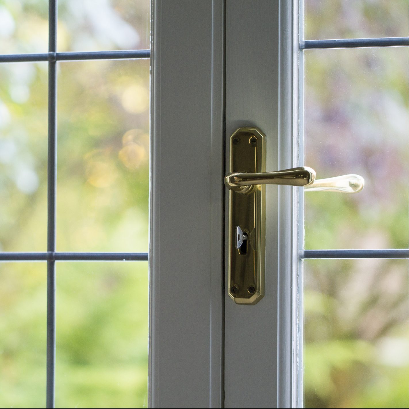 A close-up of a golden door handle on a white window, reflecting soft outdoor light and surrounded by blurred greenery.