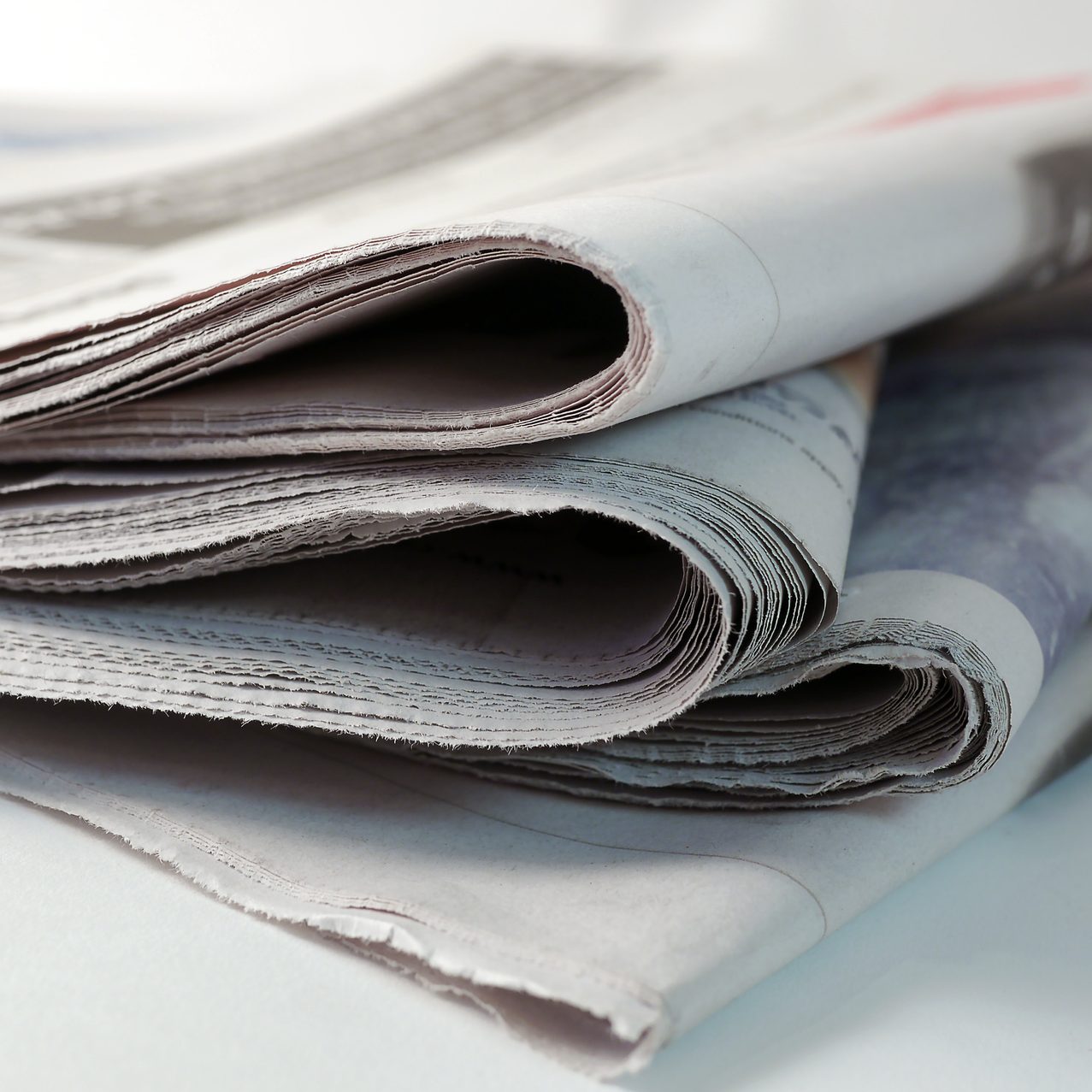 Stacked newspapers lie folded, showcasing worn edges, resting on a flat surface in a softly lit space.