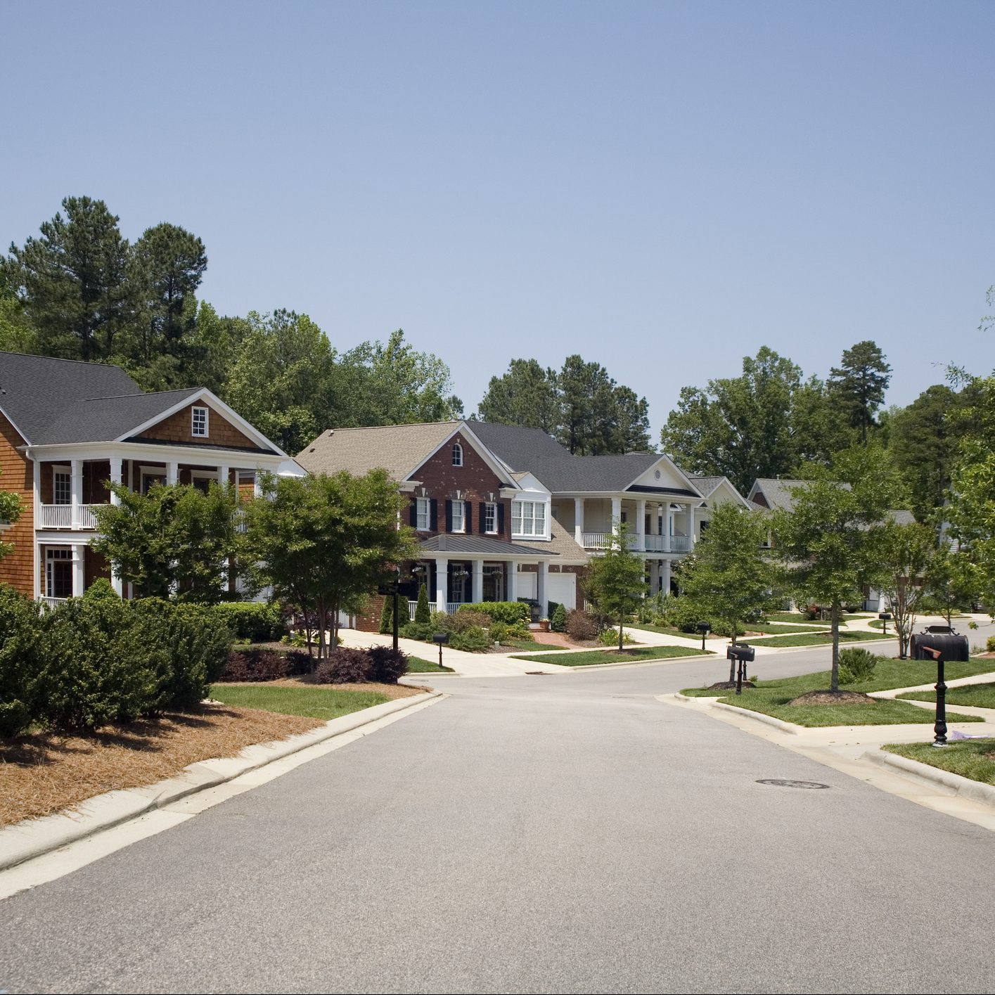 A row of suburban houses lines a quiet street, surrounded by greenery and trees under a clear blue sky.