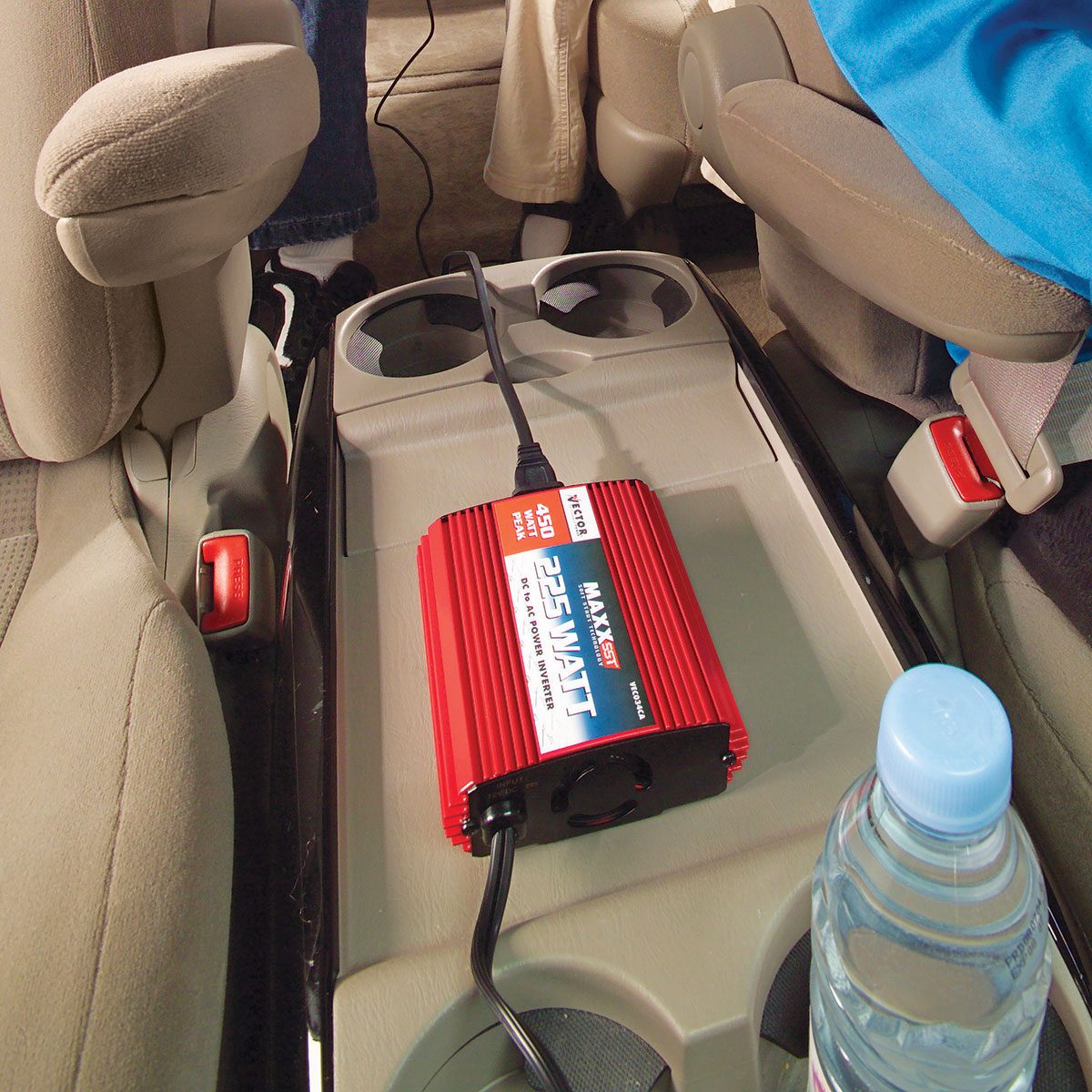 A red power inverter sits on a car's center console, connected to a power source, while a water bottle is nearby, indicating a travel context.